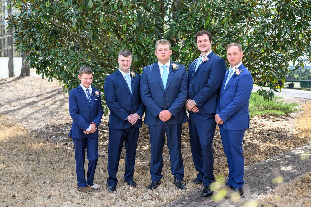 Five men in matching navy blue suits with boutonnieres pose together outdoors near a tree at a wedding.