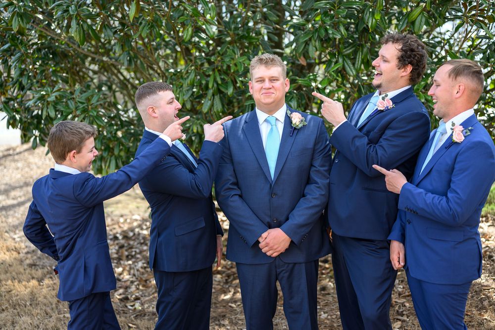 Groom in blue suit with boutonniere stands proudly as four groomsmen in navy suits point and gesture at him outdoors.