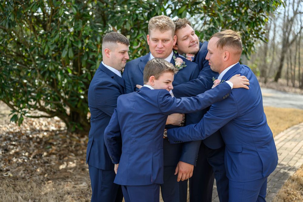 Five men in matching navy blue suits share a group hug outdoors at a wedding, surrounded by lush greenery.
