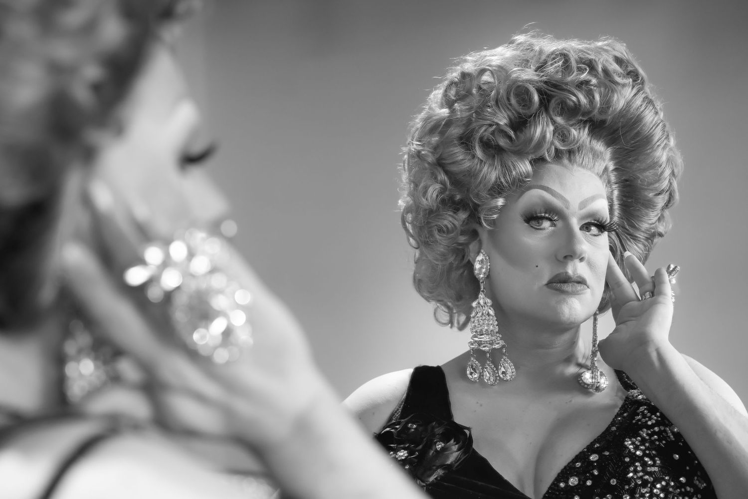 Drag performer in black sequin dress applies makeup while gazing into mirror, captured in black and white.