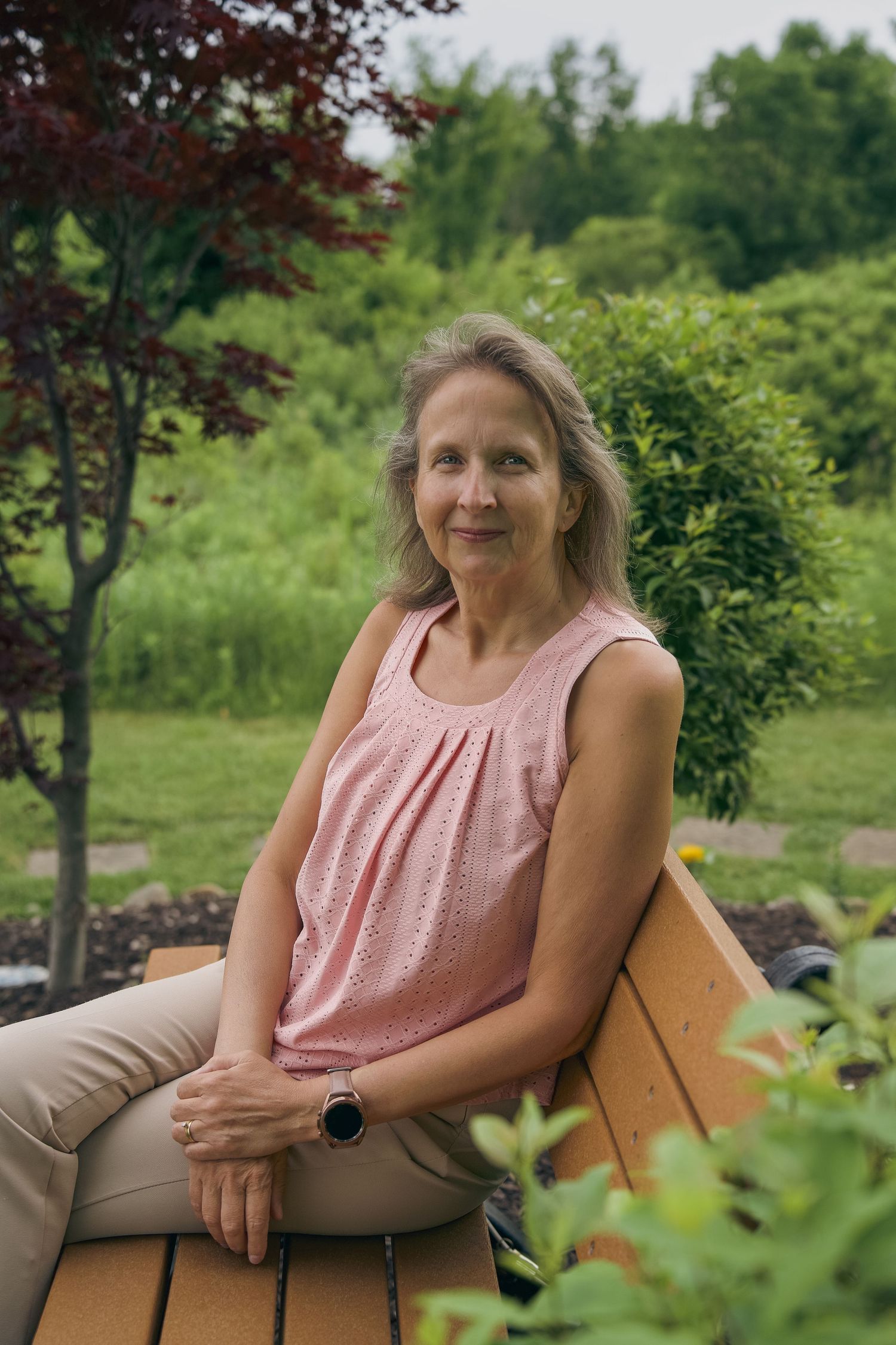 Middle-aged woman in pink sleeveless top sitting on wooden bench in lush green garden, smiling outdoors.