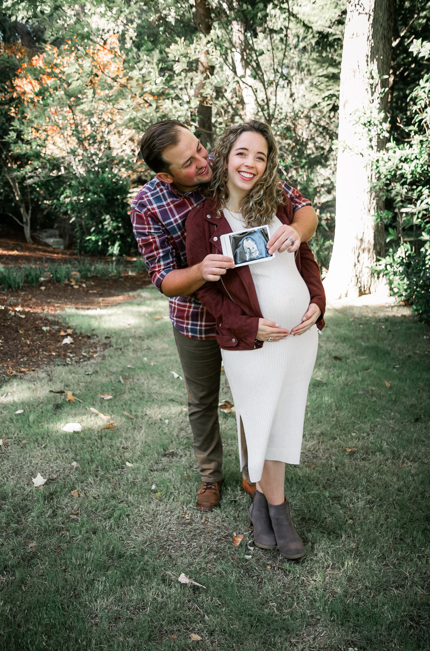 Pregnant couple smiling outdoors holding ultrasound photo, woman in white dress and man in plaid shirt.