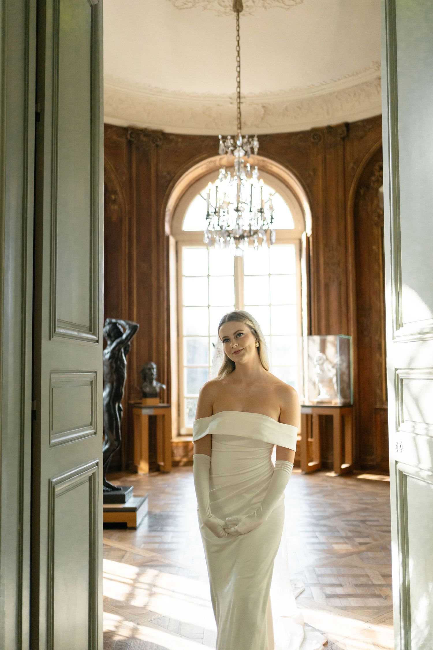 Bride in off-shoulder white gown poses in elegant wood-paneled room with crystal chandelier. Musee rodin elopement wedding