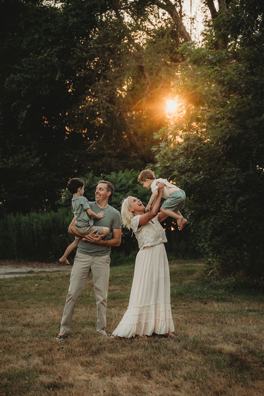 Family of four enjoying a golden sunset in a lush green park, parents holding young children in warm evening light.