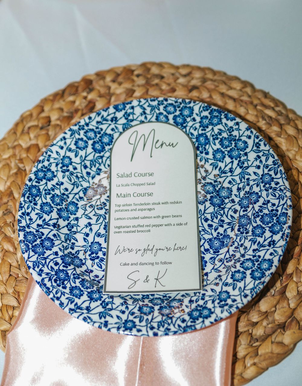 Blue floral china charger plate with elegant wedding menu card for S&K, placed on woven rattan placemat.
