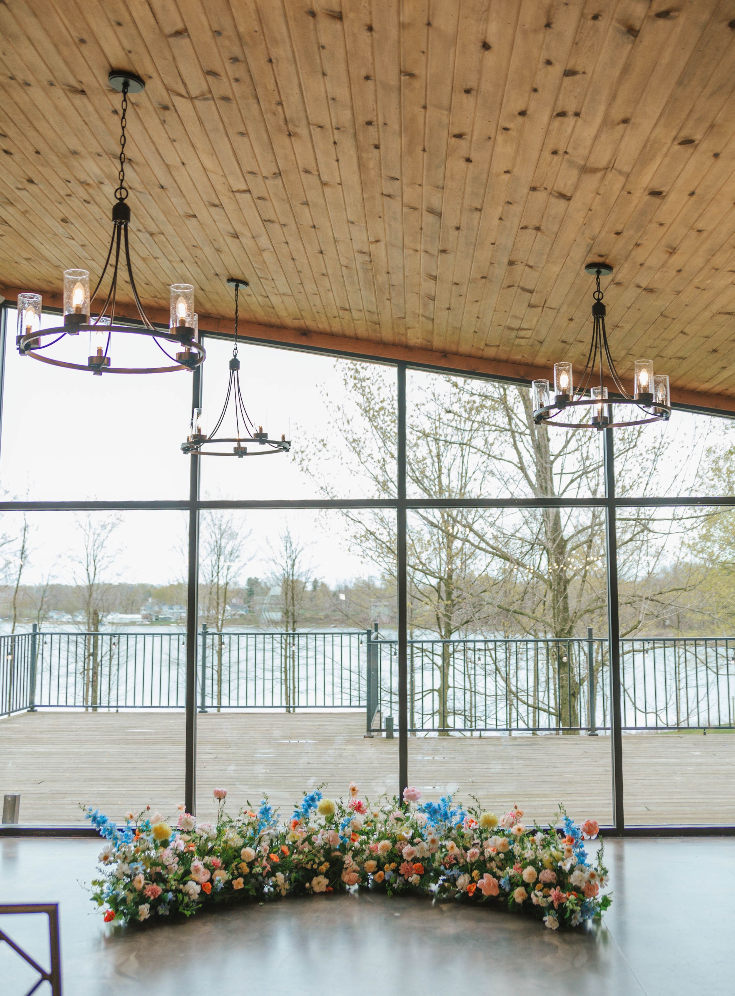 Elegant wedding venue with wood ceiling, chandelier lighting, floor-to-ceiling windows, and colorful floral arrangement on floor.