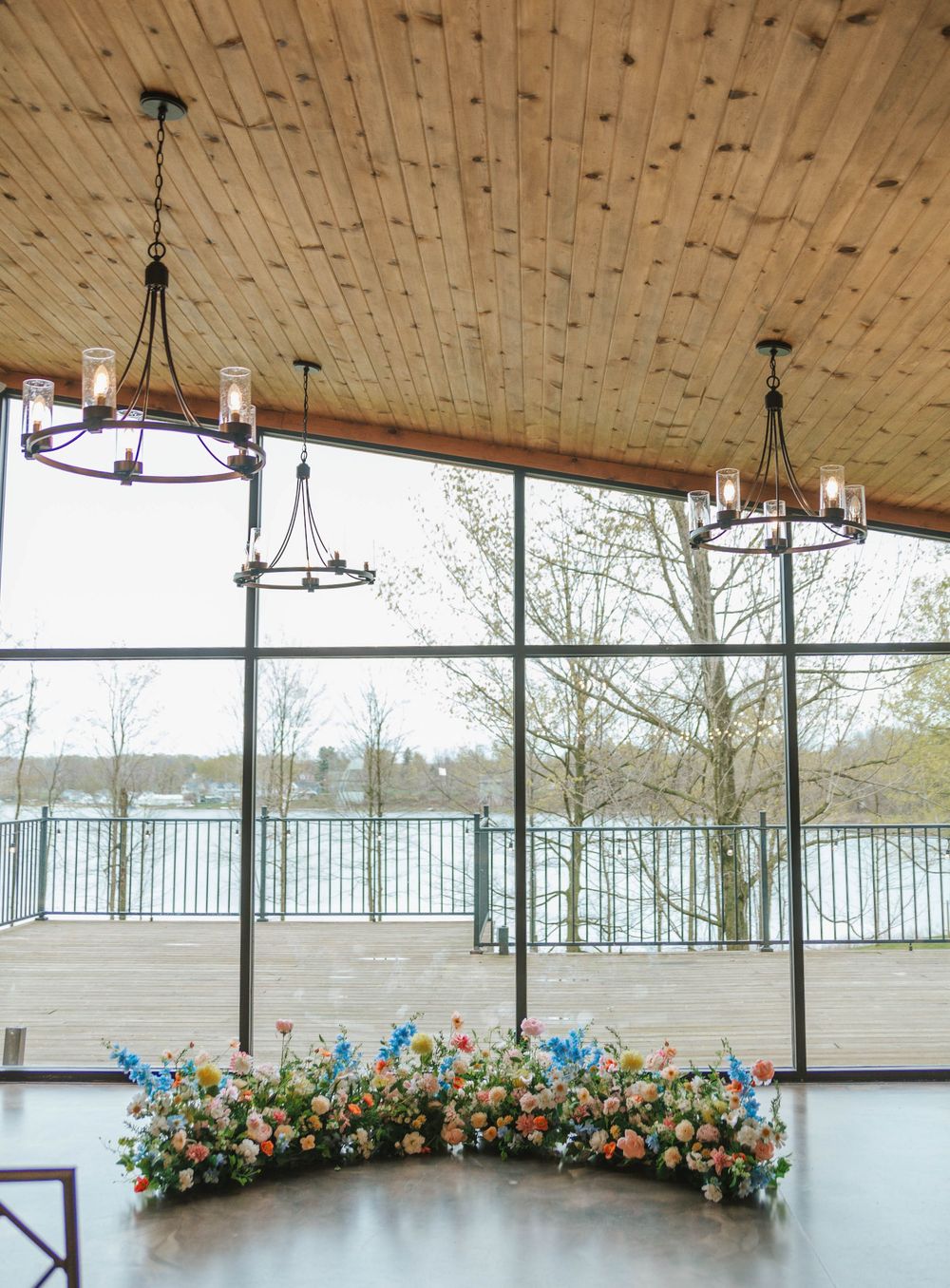Elegant wedding venue with wood ceiling, chandelier lighting, floor-to-ceiling windows, and colorful floral arrangement on floor.