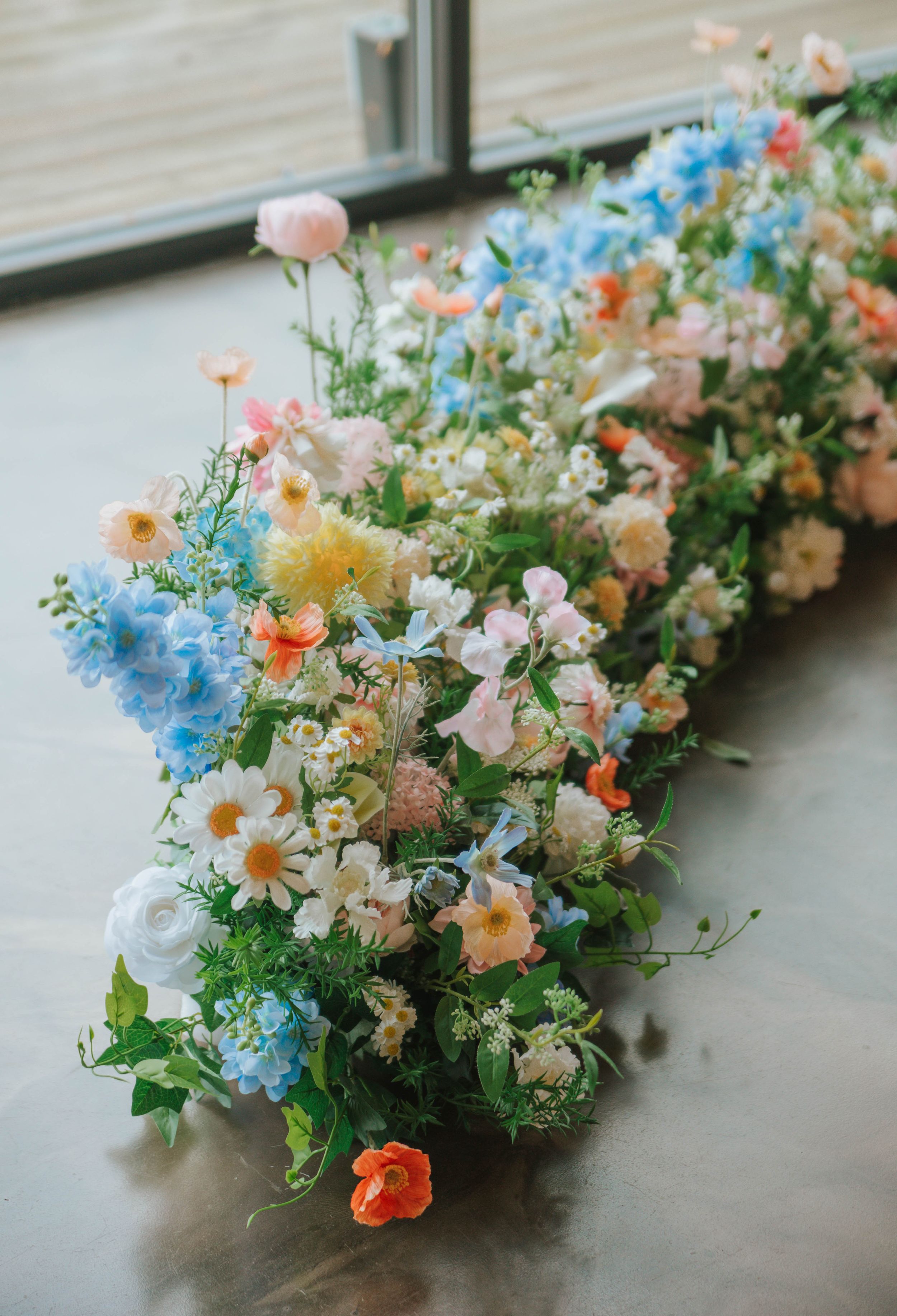 Lush floral arrangement of blue, pink, orange, and white wildflowers sprawled on a concrete floor near a window.