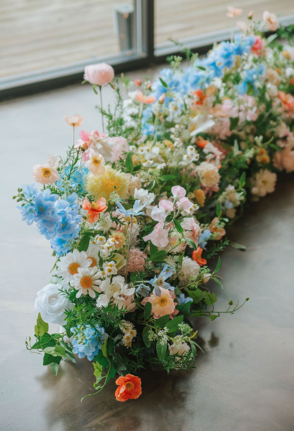 Lush floral arrangement of blue, pink, orange, and white wildflowers sprawled on a concrete floor near a window.
