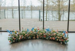 Colorful floral arrangement with pink, blue, and orange blooms displayed on a dark floor near large windows overlooking a waterfront.
