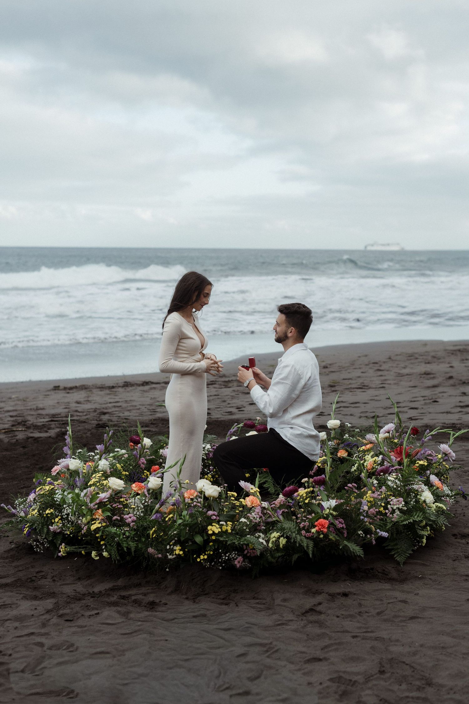 Man proposes to woman on black sand beach surrounded by a floral arrangement with ocean waves in the background.