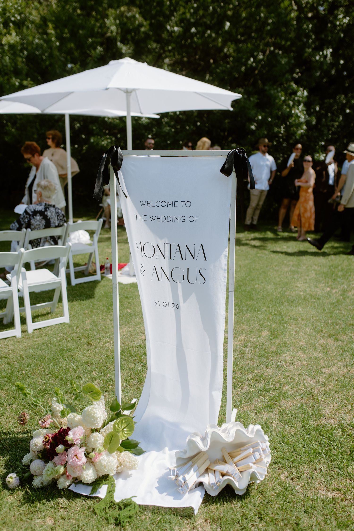 Elegant outdoor wedding welcome sign for Montana and Angus, displayed on white stand with floral arrangement and umbrella.