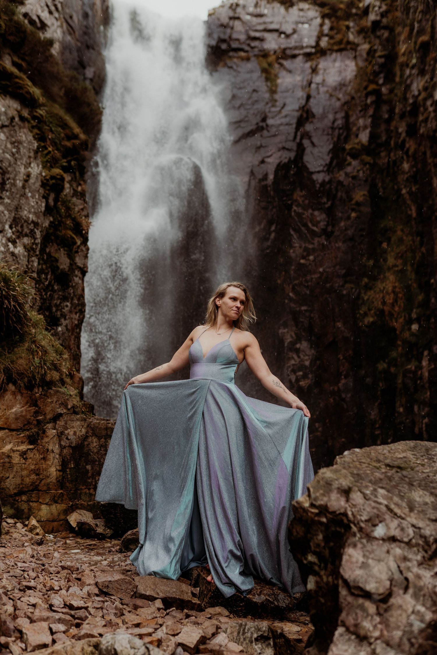 Woman in flowing blue-gray gown poses gracefully before a dramatic waterfall cascading down rocky cliffs.