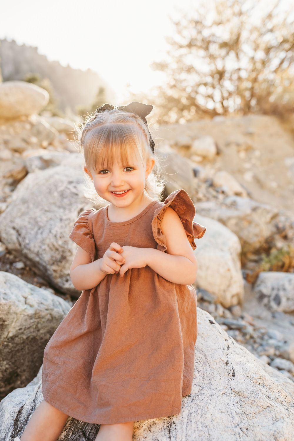 Smiling toddler girl in brown dress with bow hair ties sitting on rocks in golden sunlight outdoors.