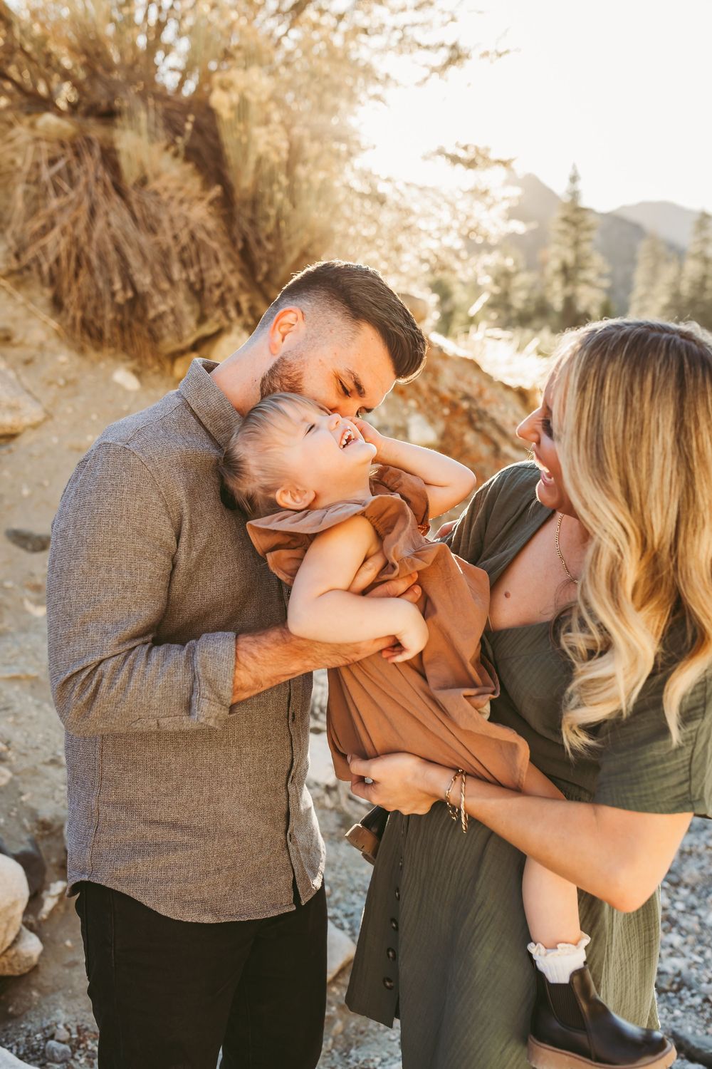 A couple shares a tender moment kissing their baby outdoors in a sun-drenched rocky landscape.