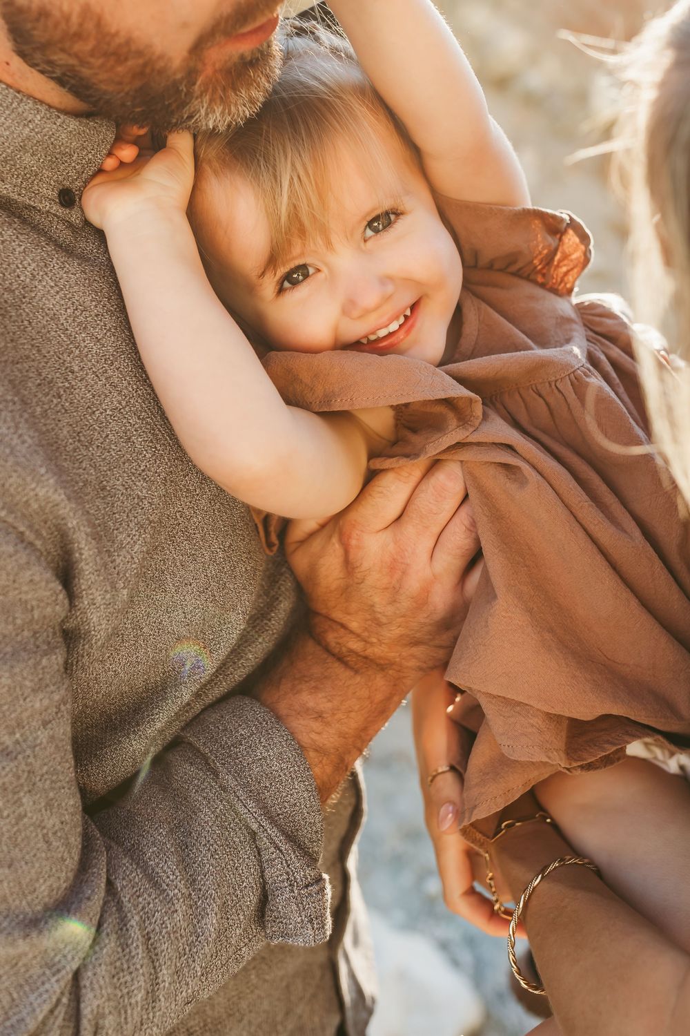 A smiling toddler girl in a dusty rose dress being held by her parents in a warm, sunlit outdoor setting.