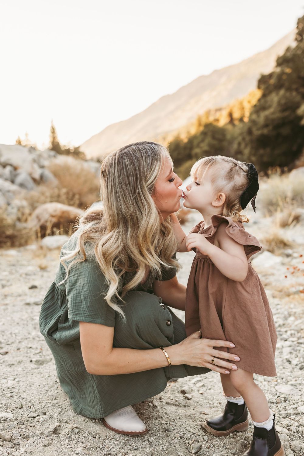 A mother crouches to kiss her toddler daughter outdoors, both wearing green and brown dresses with mountains behind them.