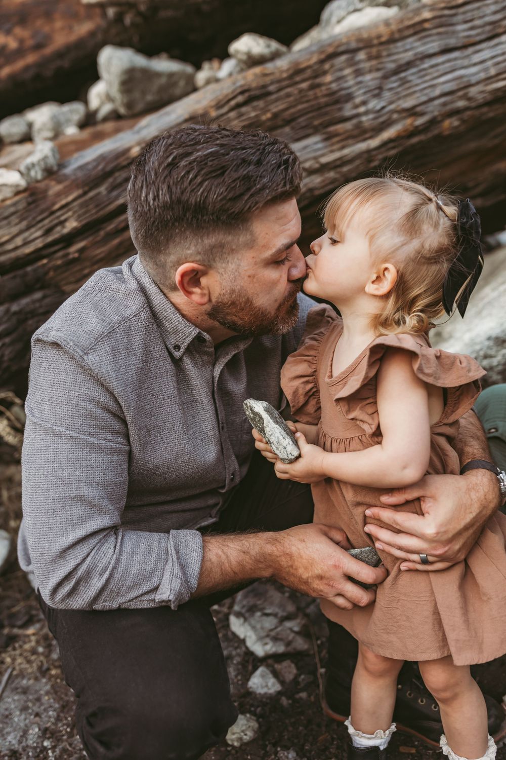 A bearded father kisses his toddler daughter outdoors near rustic logs, as she holds a rock.