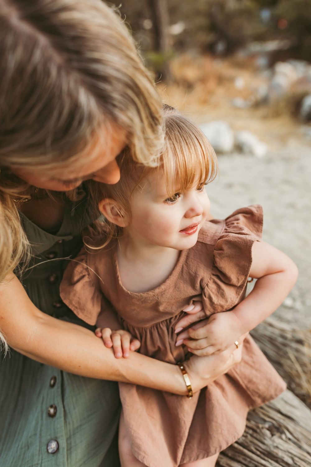A mother tenderly embraces her young toddler daughter outdoors, both wearing earthy tones in a warm, natural setting.
