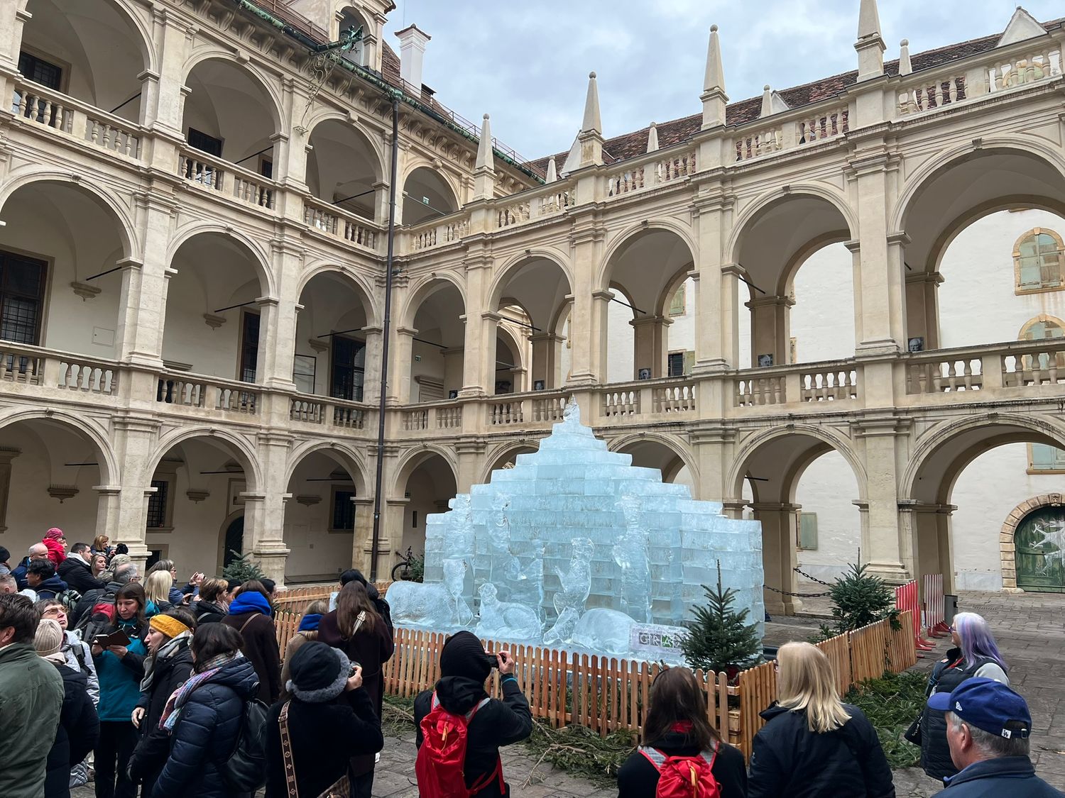 Tourists admire a large ice sculpture nativity scene in the arcaded Renaissance courtyard of a historic European palace.