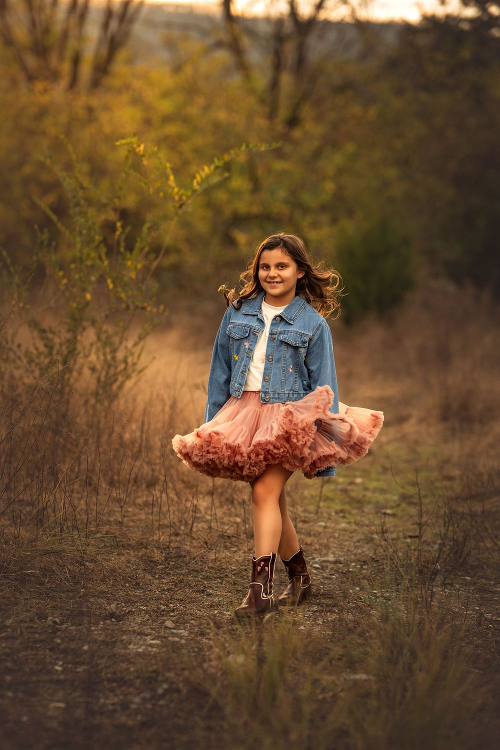 Young girl in pink tutu skirt and denim jacket twirling outdoors during golden hour for family photo session.