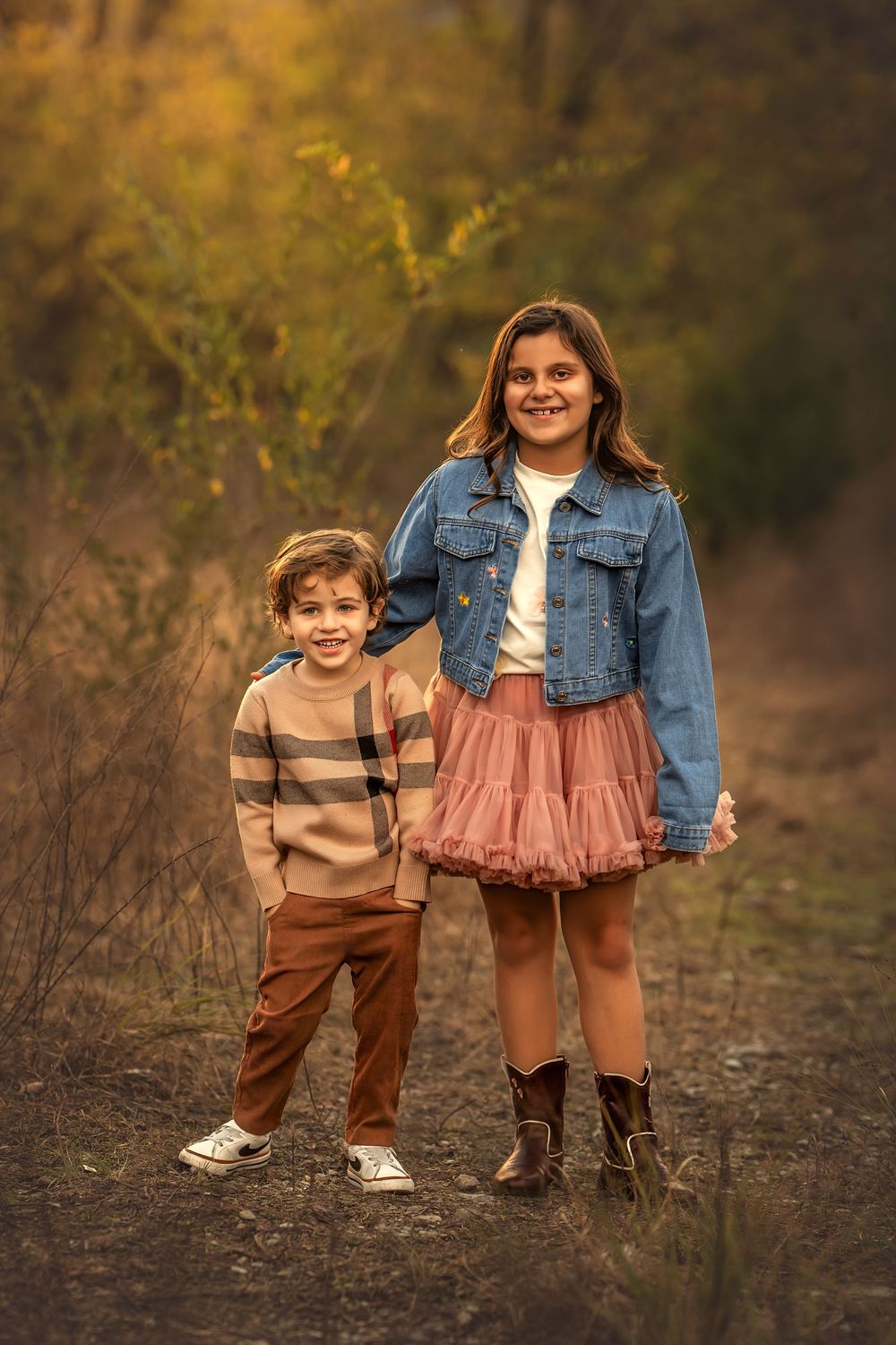 Two smiling children pose on a forest path in fall, girl in denim jacket and pink skirt, boy in striped sweater.