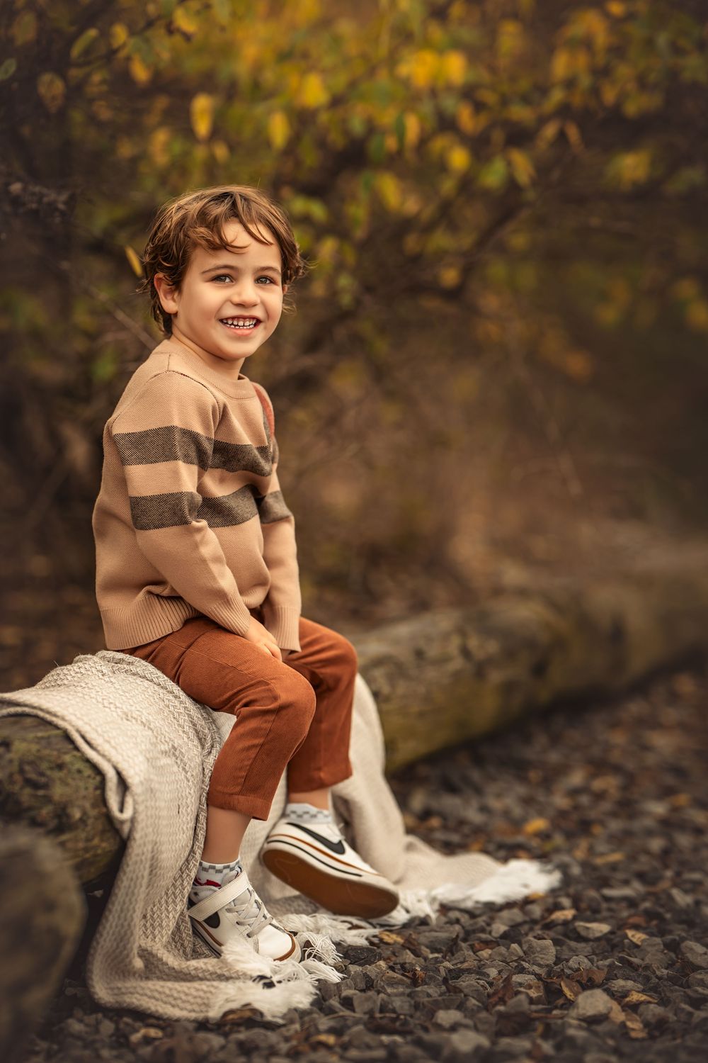 Smiling boy in striped sweater and rust pants sitting on a log in an autumn forest setting.