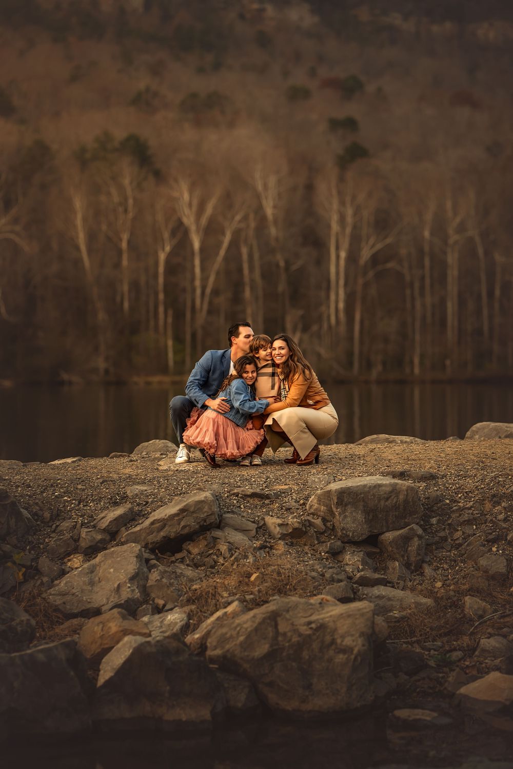 Family of four posing together on rocky lakeside shore with autumn forest backdrop for family photos with Marietta photographer.