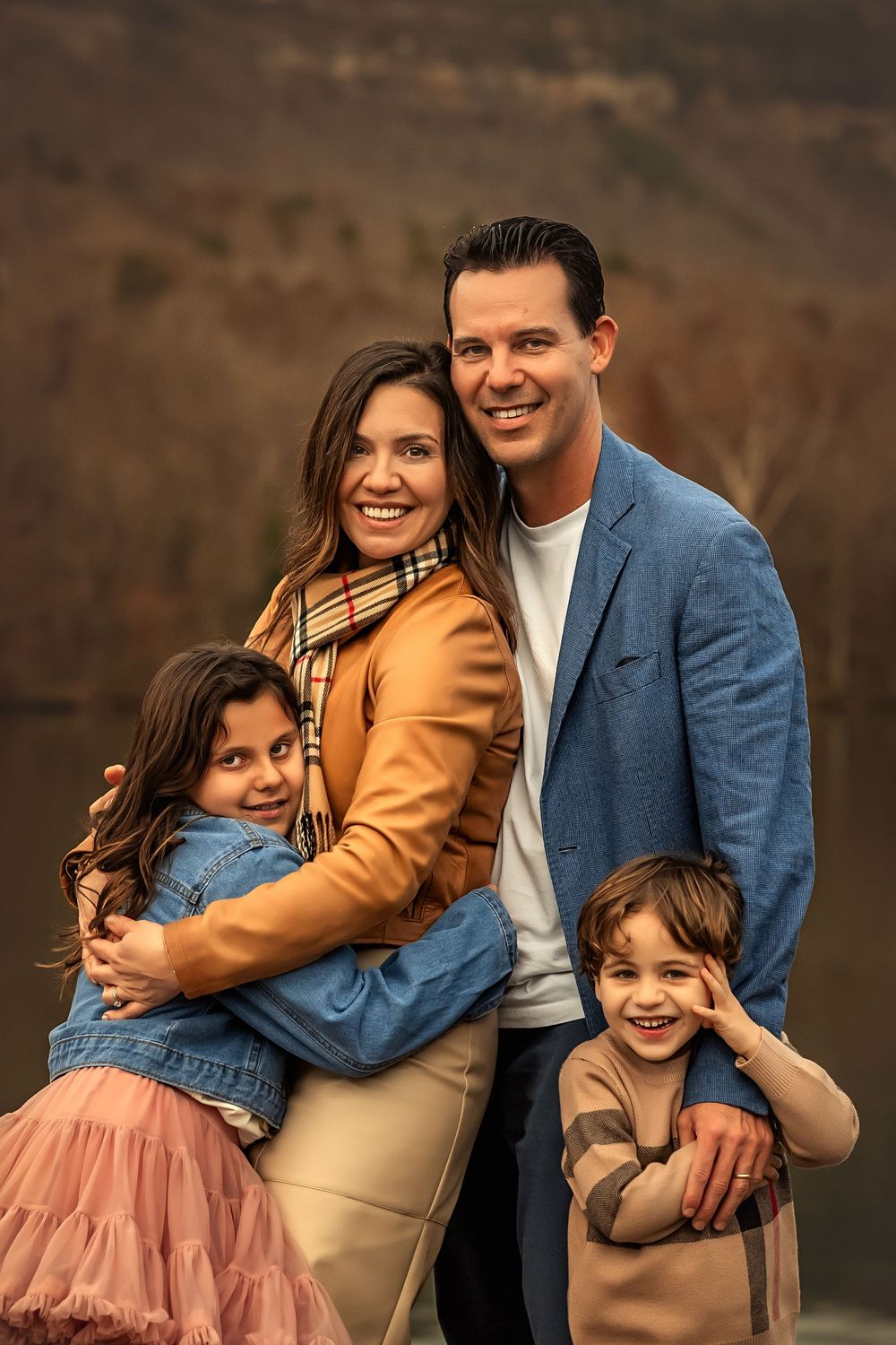 A happy family of four poses outdoors in fall attire, with parents embracing two young children near a scenic lake.