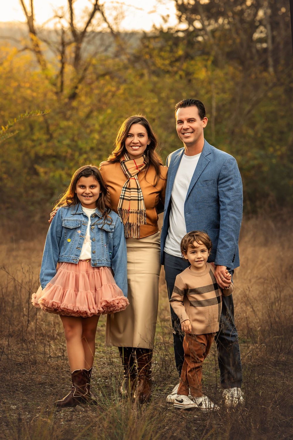 Family of four posing outdoors in fall foliage, wearing coordinated autumn outfits in warm tones of blue, gold, and brown.