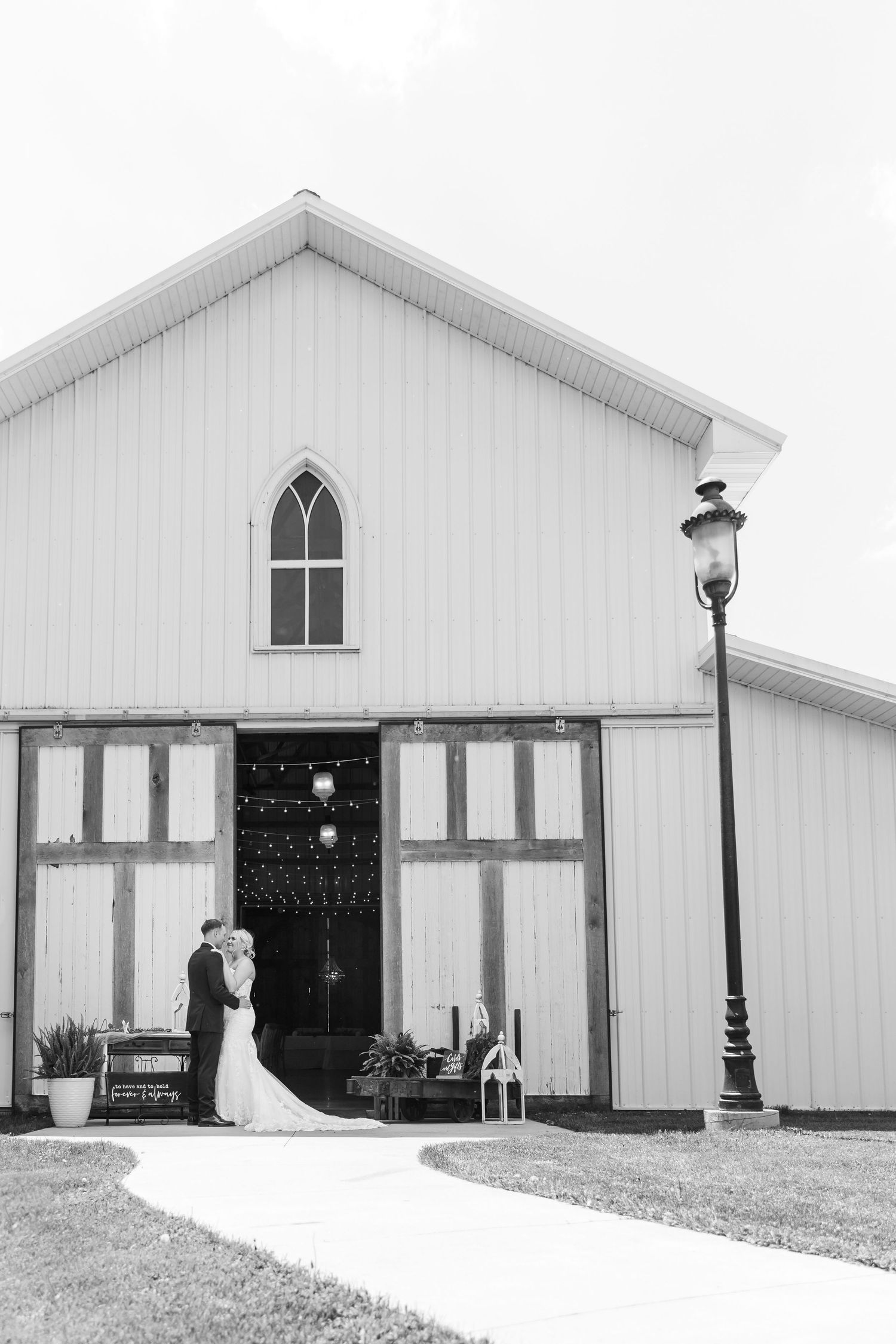 Black and white photo of a bride and groom kissing outside a white barn wedding venue with string lights.