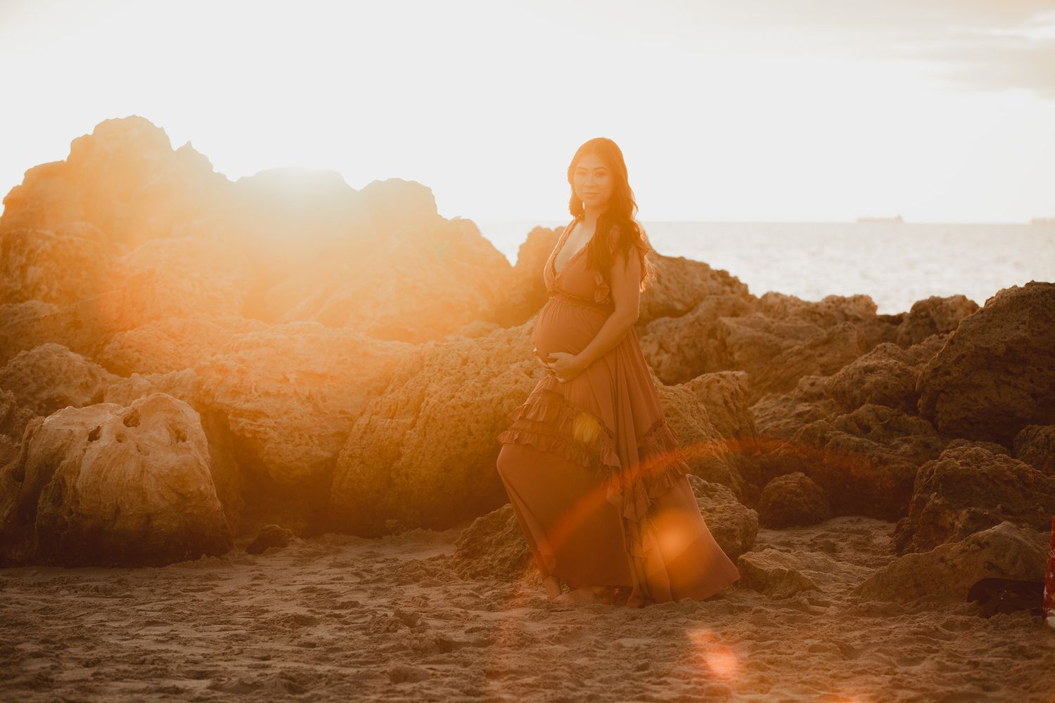Pregnant woman in flowing orange dress posing on rocky beach with dramatic golden sunset behind her.