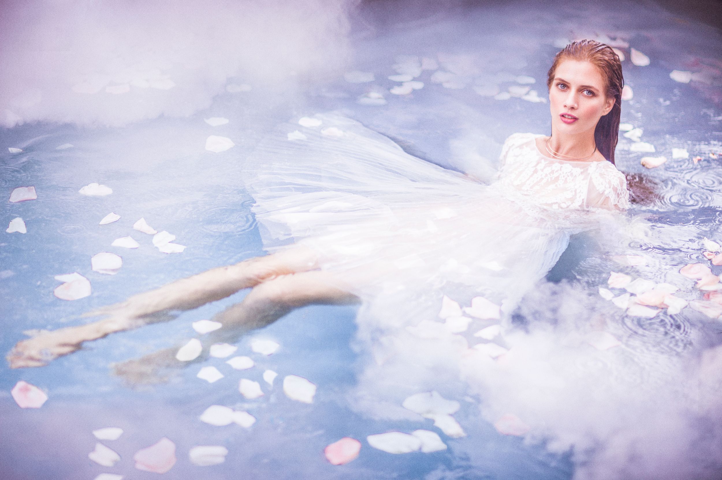 Woman in white dress floating gracefully in a misty pool surrounded by rose petals in a dreamy, ethereal setting.