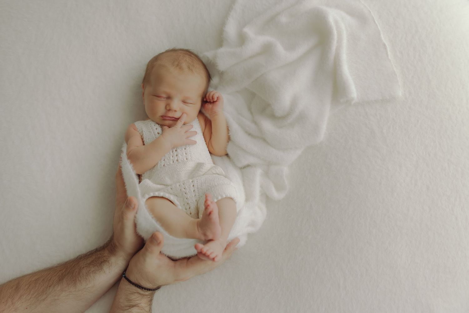 Newborn baby in white knit outfit sleeping peacefully while being gently supported by parent's hands.