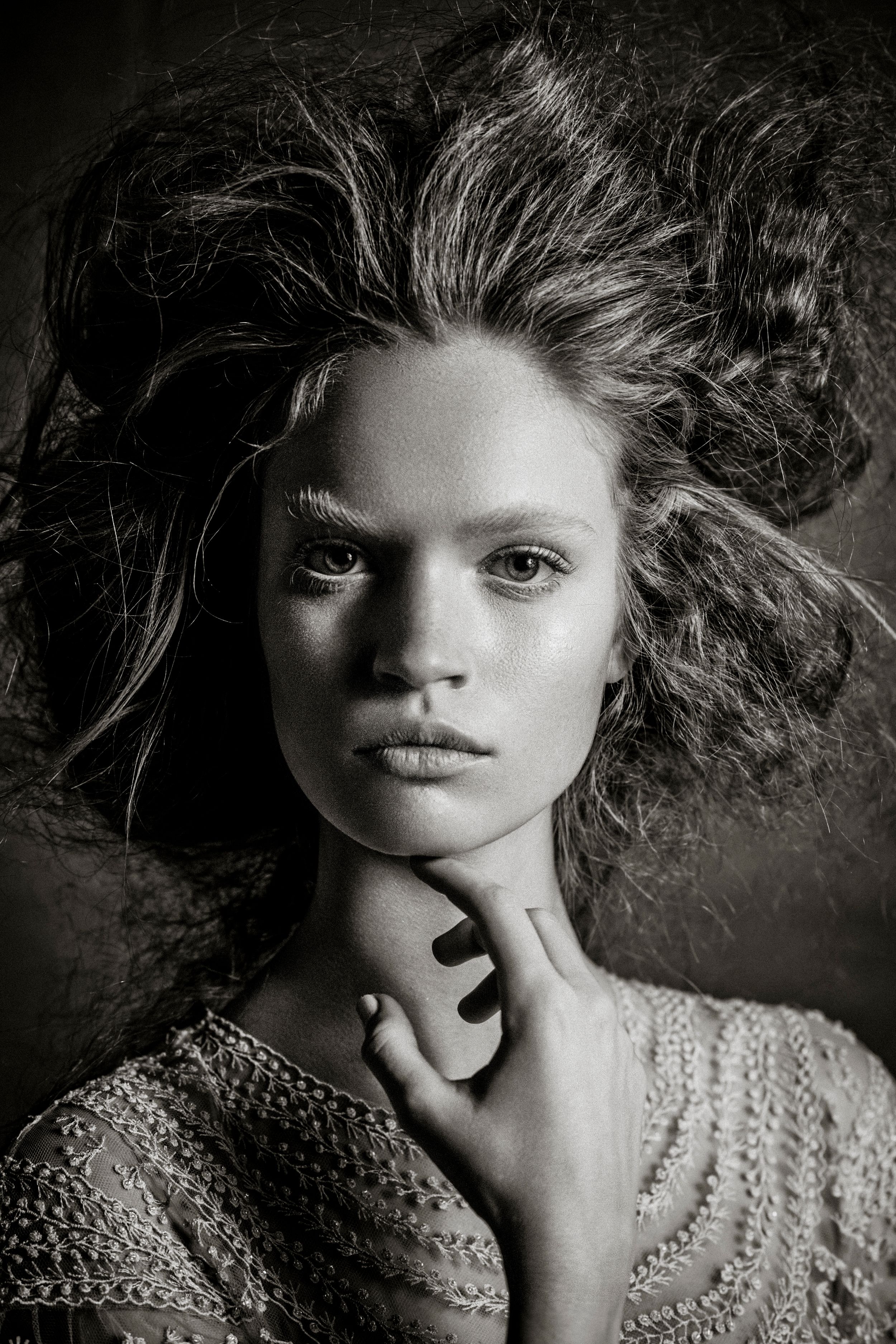 Black and white portrait of a young woman with wild flowing hair, hand on chin, wearing lace top.