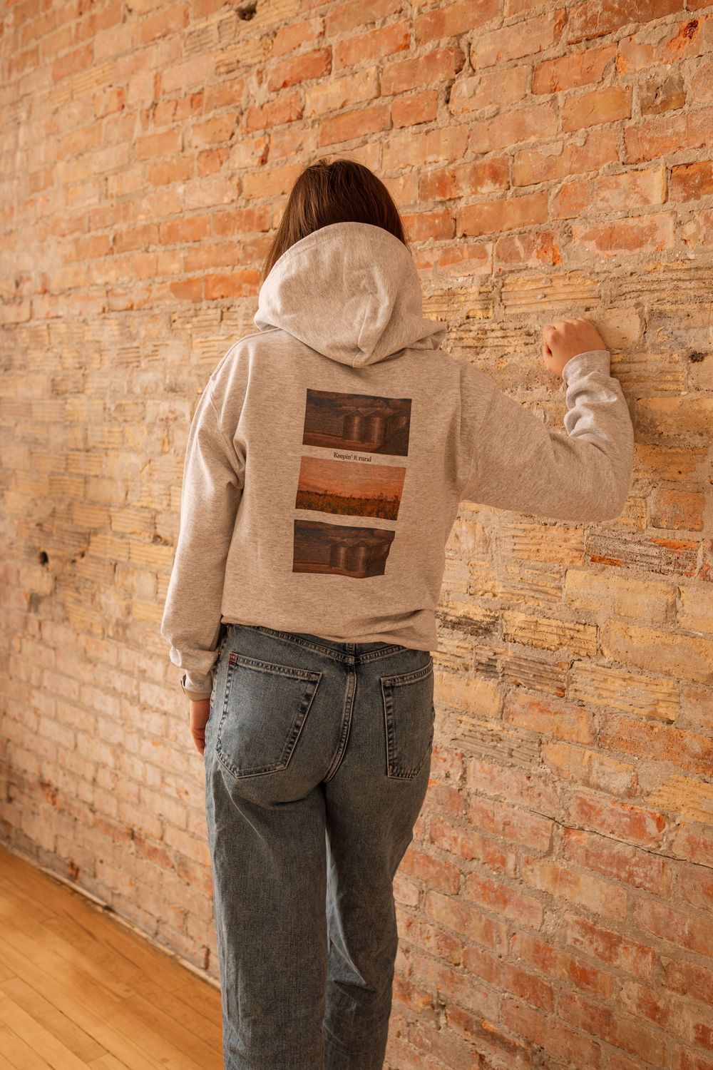 Woman wearing a gray graphic hoodie and jeans, shown from behind against a rustic brick wall.