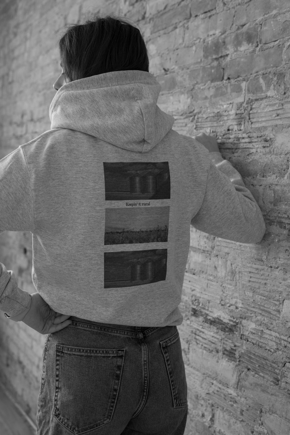 Black and white photo of person wearing gray hoodie with graphic print on back, standing against brick wall.