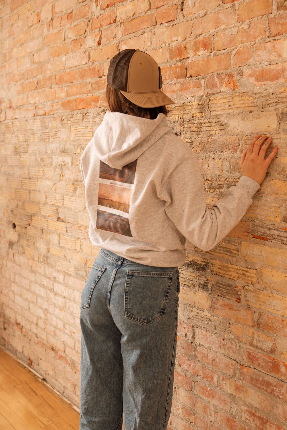 Person wearing gray hoodie with back graphic, baseball cap, and jeans facing brick wall.