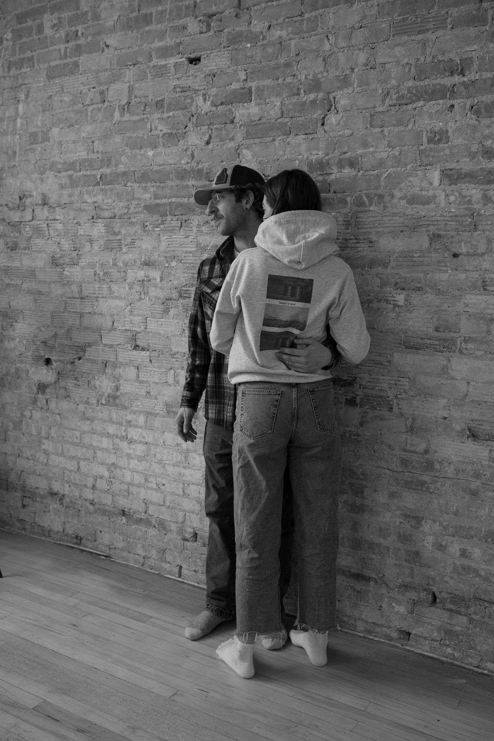 Couple in casual hoodies and jeans standing together against rustic brick wall, black and white photo.