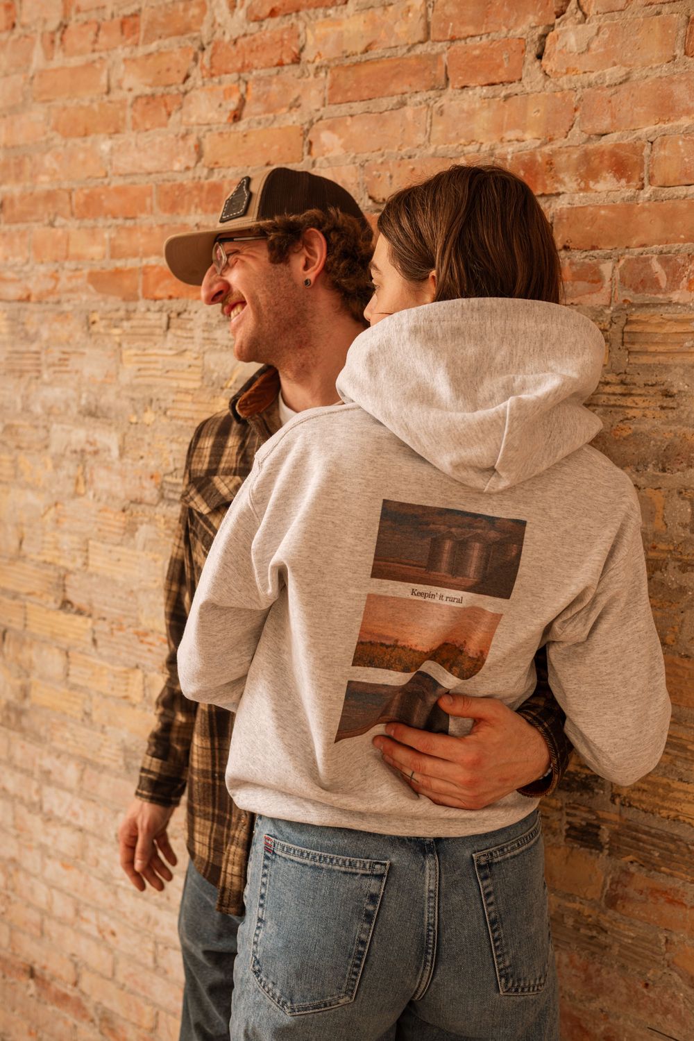Smiling couple embracing against rustic brick wall, man in plaid shirt and cap, woman in graphic back hoodie and jeans.