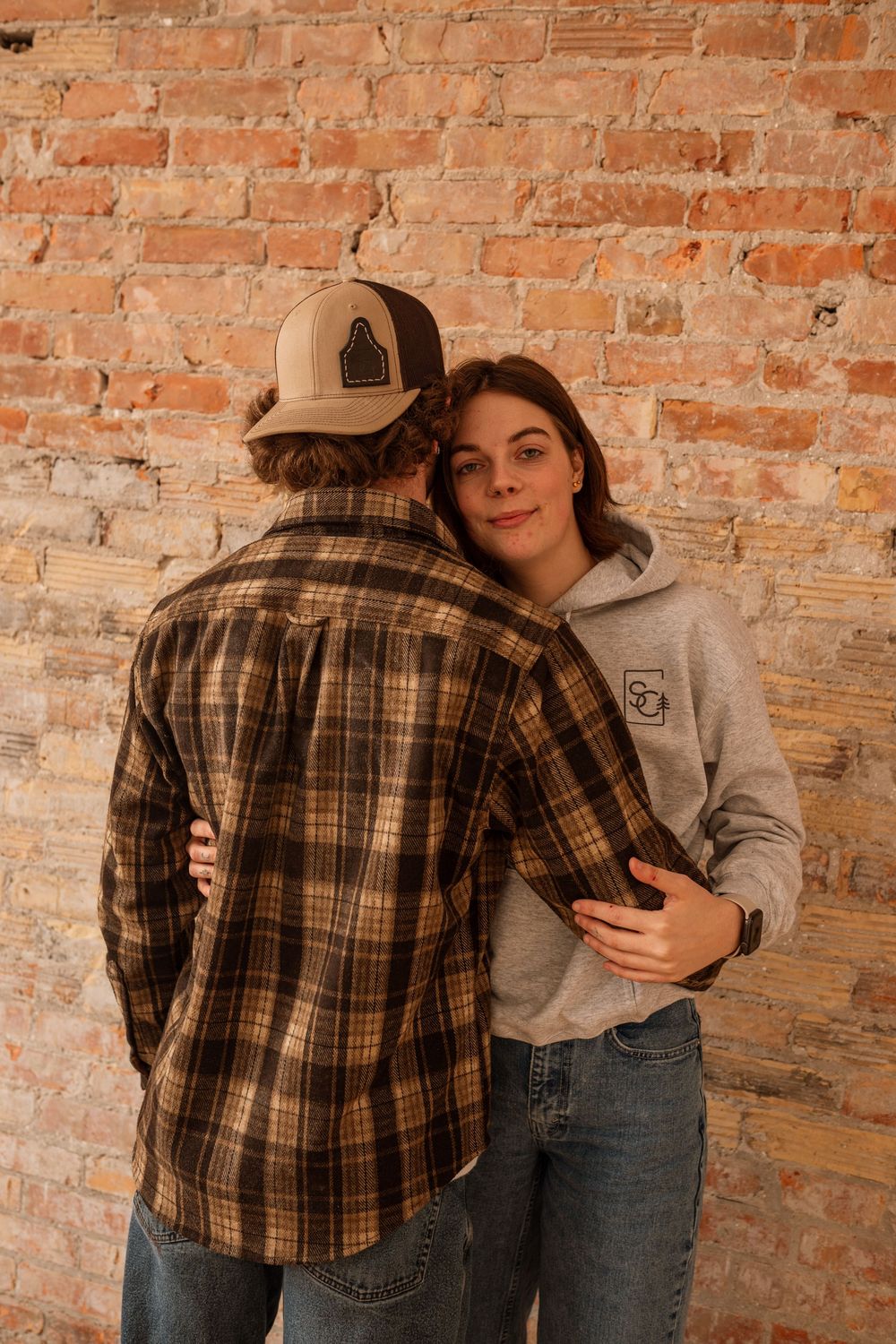 Couple embracing against rustic brick wall, man in plaid flannel shirt and cap, woman in gray hoodie and jeans.