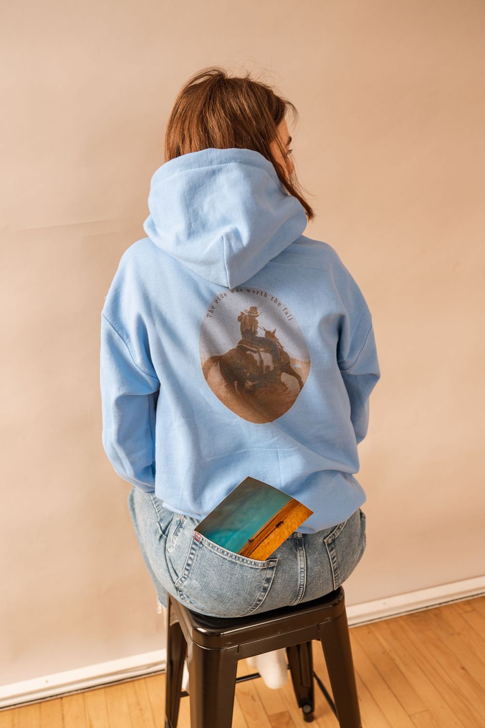 Woman sitting on stool wearing light blue graphic hoodie and jeans, shown from behind with a book in her back pocket.