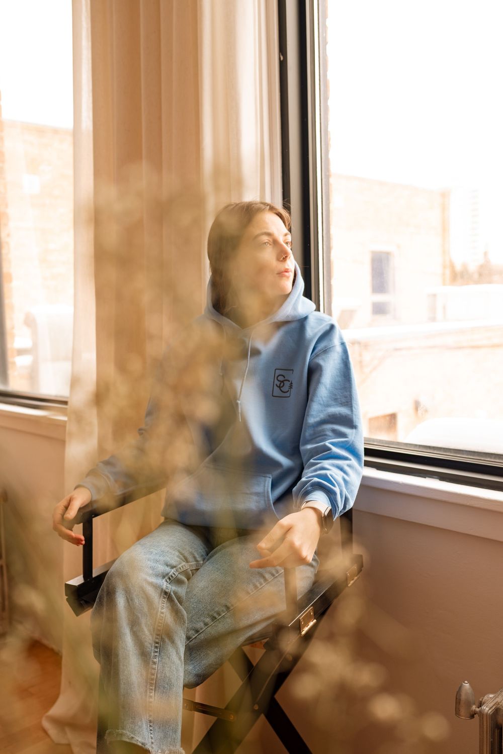 Young woman in blue hoodie sitting by sunlit window gazing outside through sheer curtains.