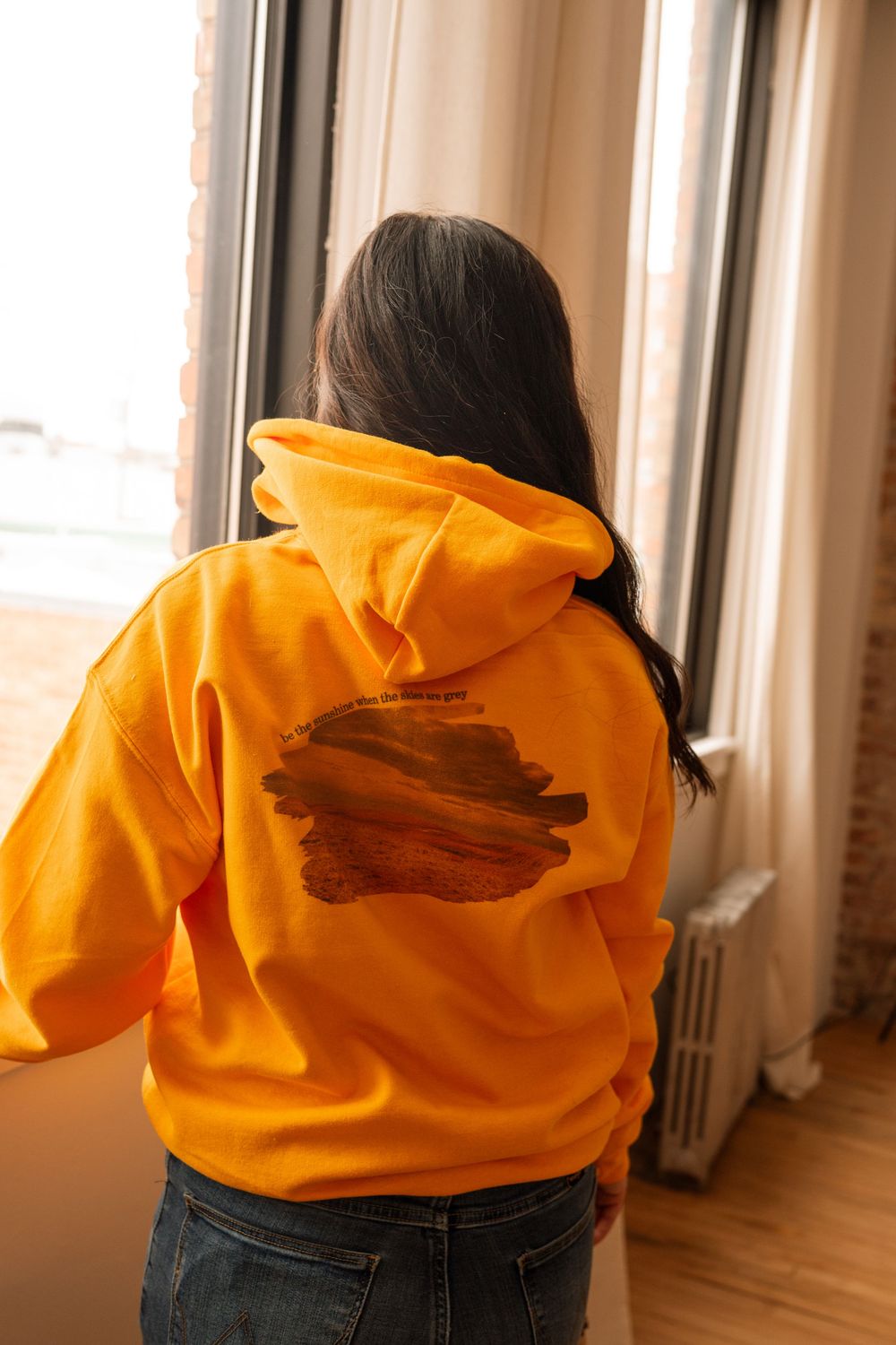 Woman in bright yellow graphic hoodie standing by window with natural light, rear view, wearing jeans.