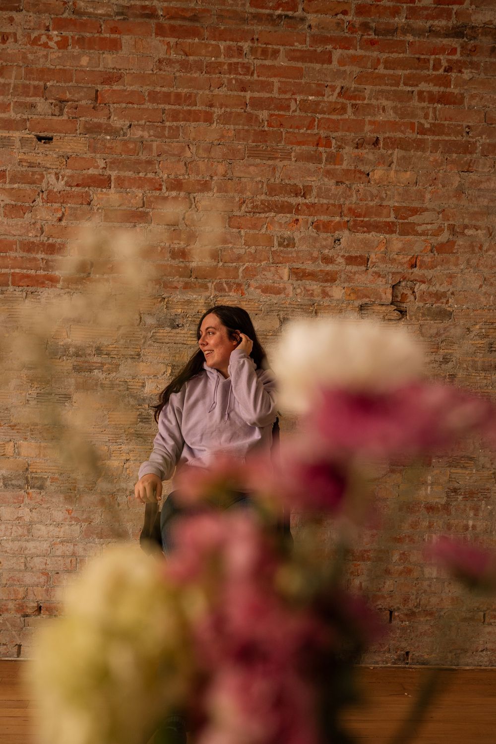 Young woman in lavender hoodie sitting against brick wall with blurred pink and white flowers in foreground.