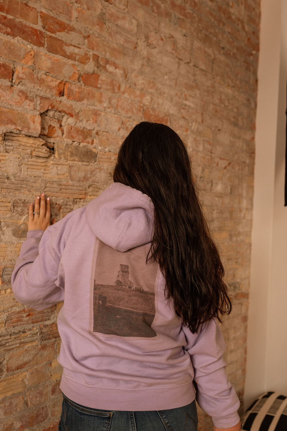 Woman in lavender graphic hoodie facing brick wall, showing back print design, wearing jeans.