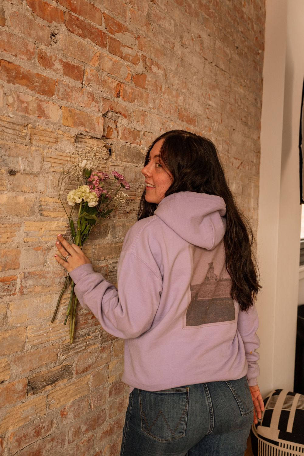 Smiling woman in lavender hoodie holding wildflowers against exposed brick wall.