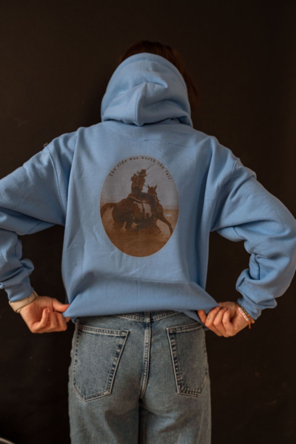 Back view of light blue graphic hoodie with western cowboy print, paired with light wash denim jeans.