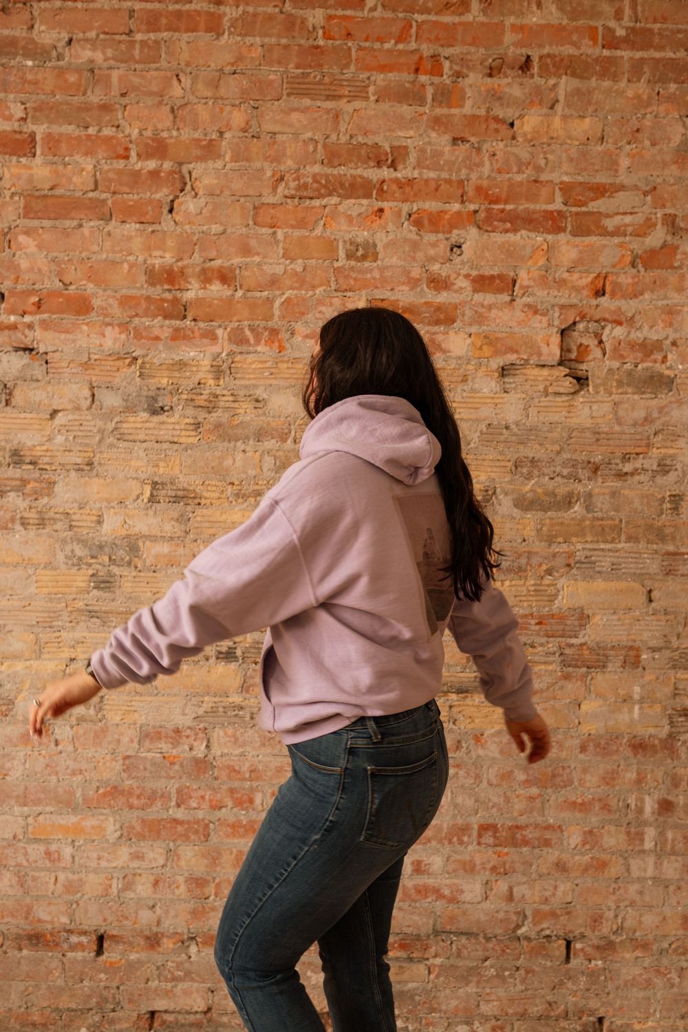 Woman dancing in front of brick wall wearing lavender hoodie and blue jeans, photographed from behind.