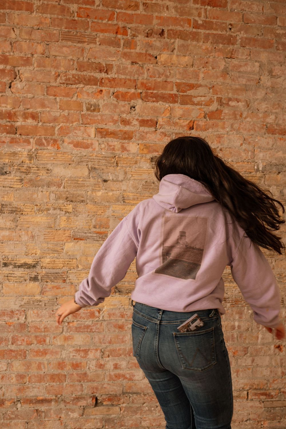 Woman in lavender graphic hoodie dancing in front of rustic brick wall, long dark hair flowing, wearing jeans.